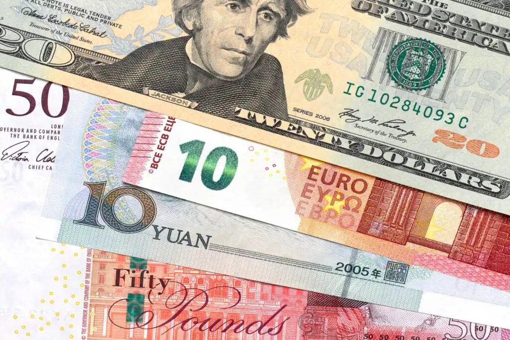 A close-up of five banknotes from different currencies: US dollar, euro, Chinese yuan, and British pound, arranged in a fan pattern, showing portions of their designs and denominations.