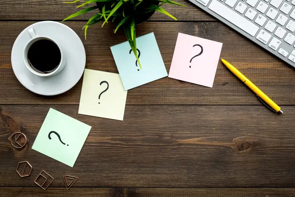 A coffee cup, a yellow pen, a keyboard, a potted plant, and four sticky notes with question marks are arranged on a wooden desk.