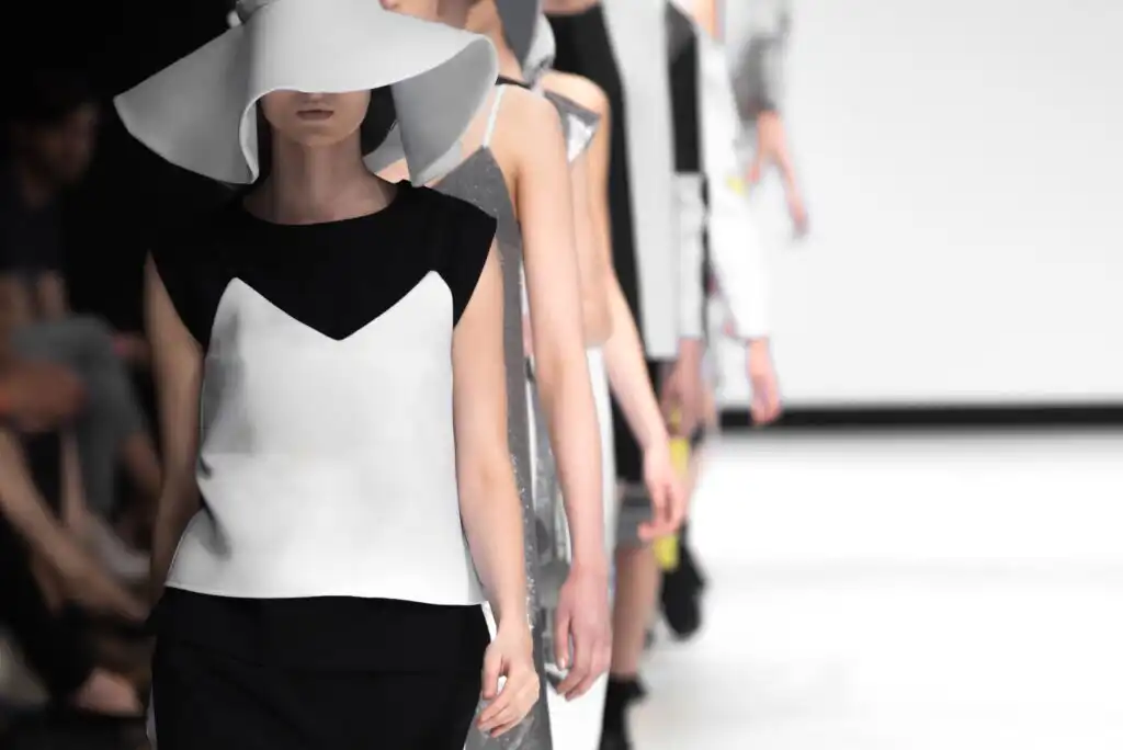 Models walk down a runway wearing modern, minimalist outfits in black, white, and silver tones. The front model wears a large white hat that shades her face and a black-and-white top. The background is bright and out of focus.