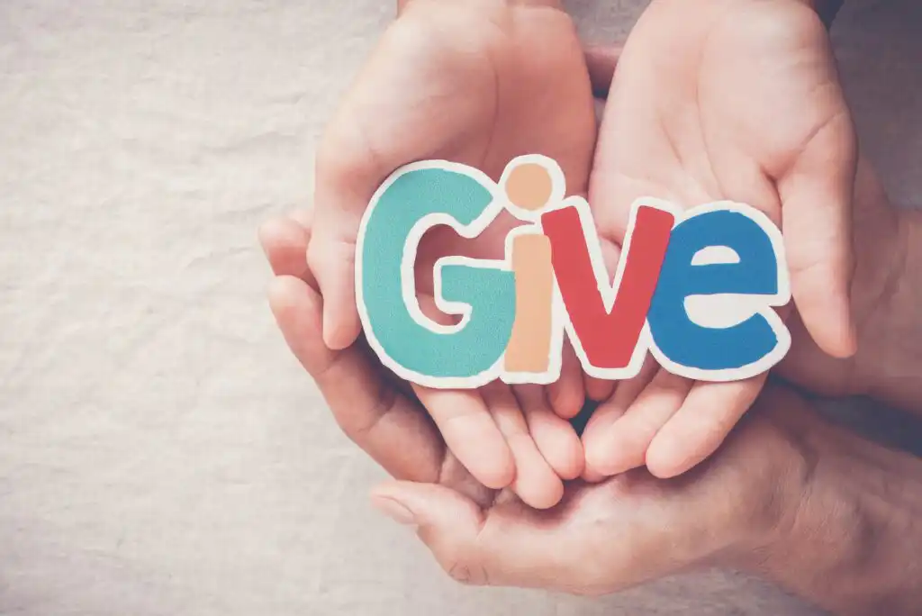 Two pairs of hands, one larger and one smaller, gently hold a colorful cutout of the word Give on a soft, neutral background, symbolizing kindness, support, and generosity.