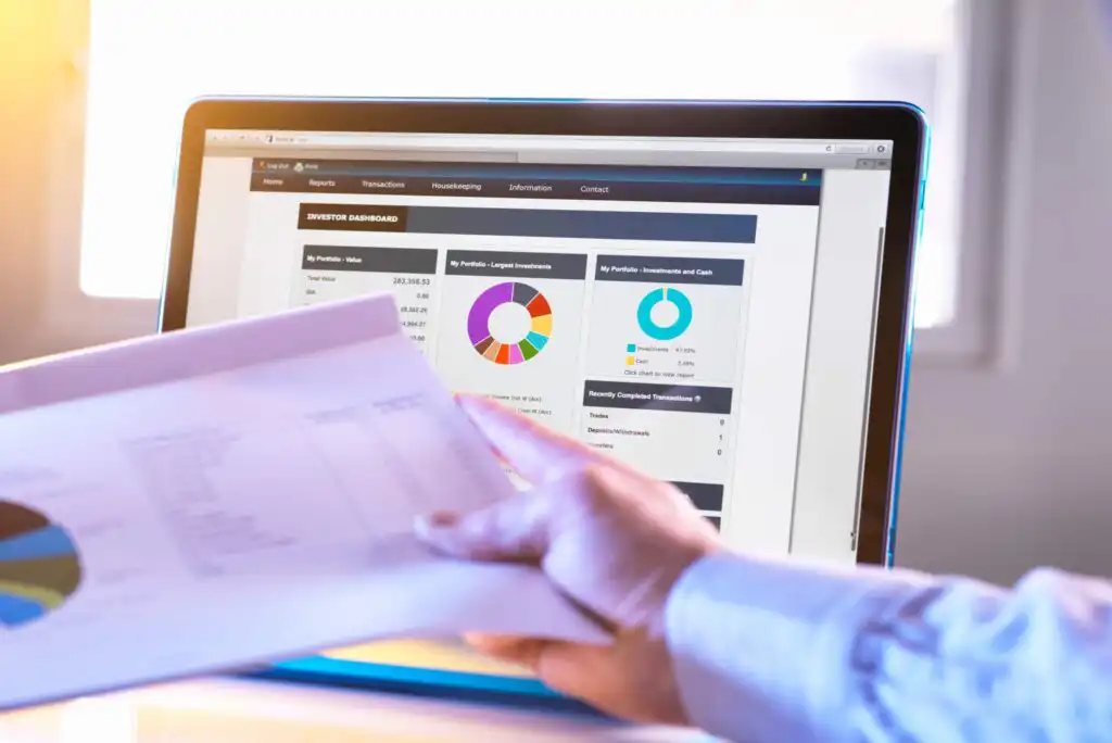A person holds a financial report in front of a computer screen displaying an investment dashboard with charts, graphs, and financial data in a bright, sunlit office setting.