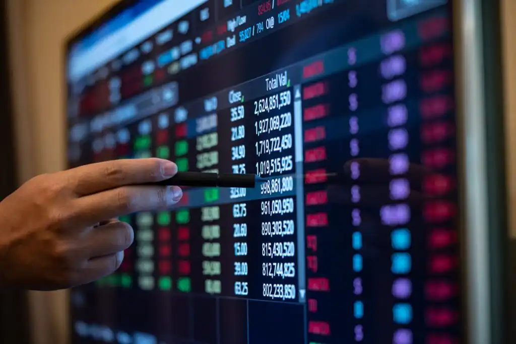 A hand holding a pen points at a large digital screen displaying rows of colorful stock market data, with numbers and figures in red, green, blue, and white.