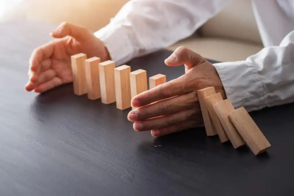 A person’s hands stop a row of falling wooden domino blocks on a dark table, preventing the rest from toppling over. The person is wearing a white long-sleeved shirt.
