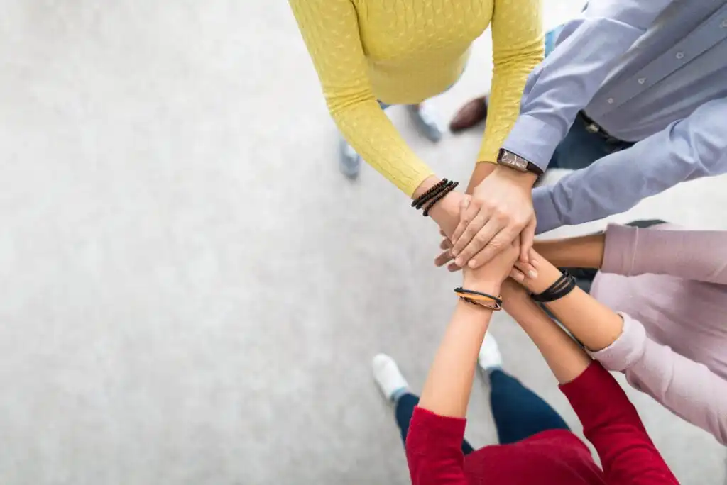 A group of people stand in a circle, stacking their hands together in the center, symbolizing teamwork and unity. The image is taken from above, showing only their arms and torsos.