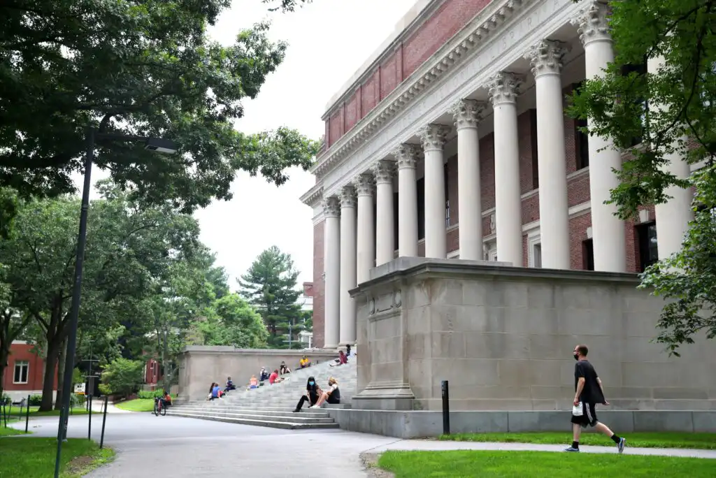 People sit and walk near the steps of a large, classical-style building with tall columns, surrounded by trees and green lawns on a college campus.
