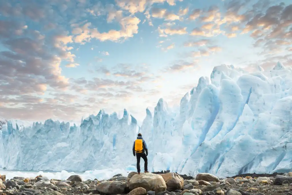 A person with a yellow backpack stands on rocky ground, facing large, jagged ice formations under a colorful sky with scattered clouds at sunrise or sunset.