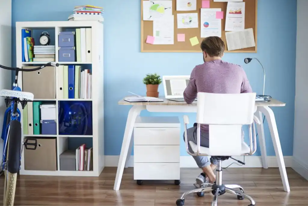 A person sits at a modern desk working on a laptop in a bright home office with blue walls, a white chair, shelves with books and binders, a corkboard with papers, and part of a bicycle visible.