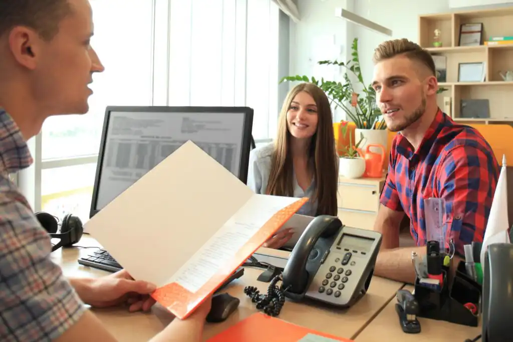 A man holding a folder talks to a smiling couple across a desk in an office, with a computer, phone, and office supplies visible on the desk.