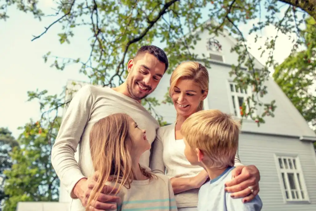 A smiling family of four stands closely together outdoors in front of a white house, surrounded by green trees. The parents look down lovingly at their two young children, who are looking up at them.