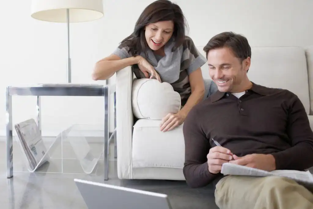 A woman and a man sit together in a bright living room, smiling as they look at a laptop. The man holds a pen and notebook, while the woman leans on a white sofa next to a glass side table with magazines.