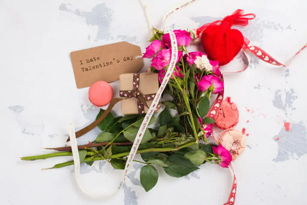 A bouquet of pink roses, broken macarons, a small wrapped gift, and a tag reading I hate Valentines Day are arranged on a white surface with decorative ribbon.