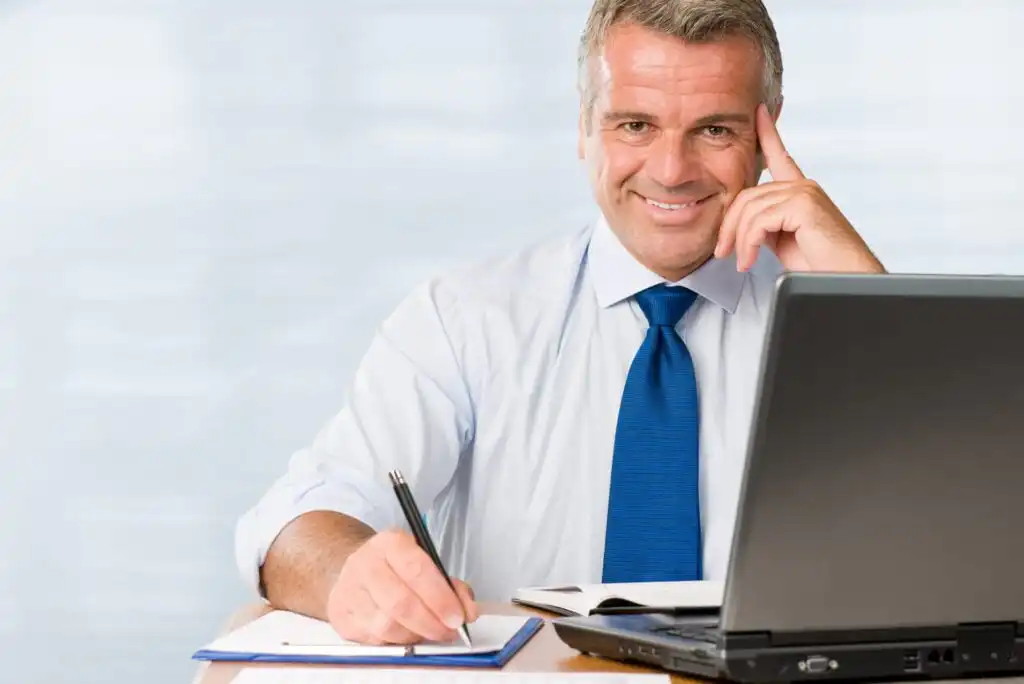 A smiling man in a blue tie sits at a desk with a laptop, writing on a notepad with a pen. An open book is also on the desk. He is looking at the camera, resting his head on one hand.