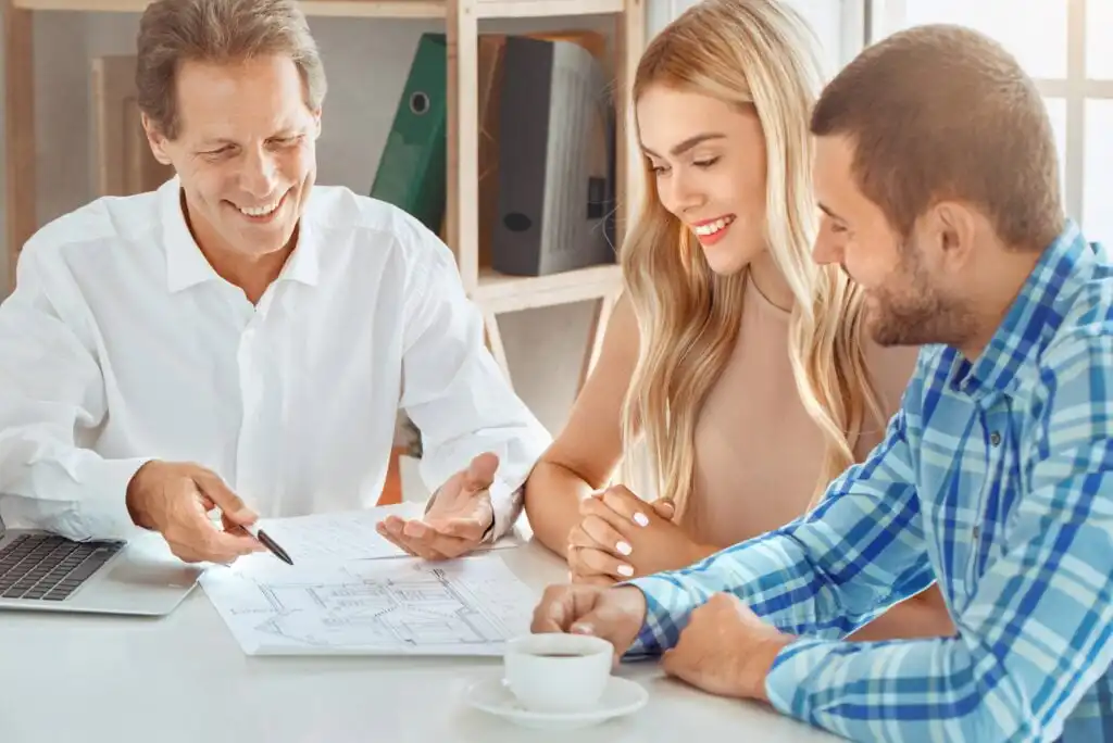 Three people sit at a table discussing documents. An older man points at a paper with architectural plans while a young woman and man listen and smile. A laptop and coffee cup are on the table.