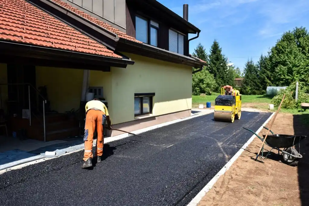 A worker levels fresh asphalt on a driveway next to a yellow house, while a yellow steamroller compacts the surface. Construction tools and a wheelbarrow are visible nearby, with trees in the background.