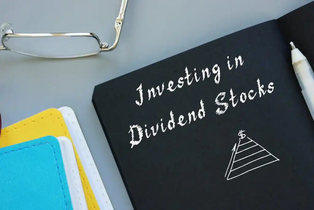 A black notebook with Investing in Dividend Stocks written in white, next to a simple pyramid diagram, a pen, eyeglasses, and two closed notebooks in yellow and blue.