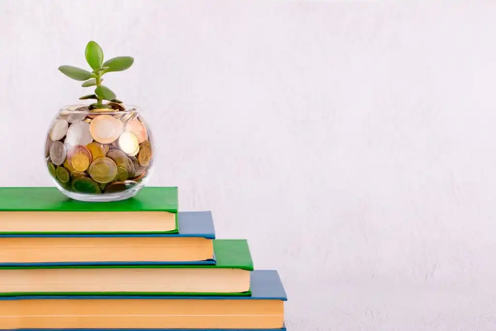 A small plant in a glass jar filled with coins sits on top of a stack of four books with green and blue covers, symbolizing growth, investment, and education. The background is plain and light-colored.