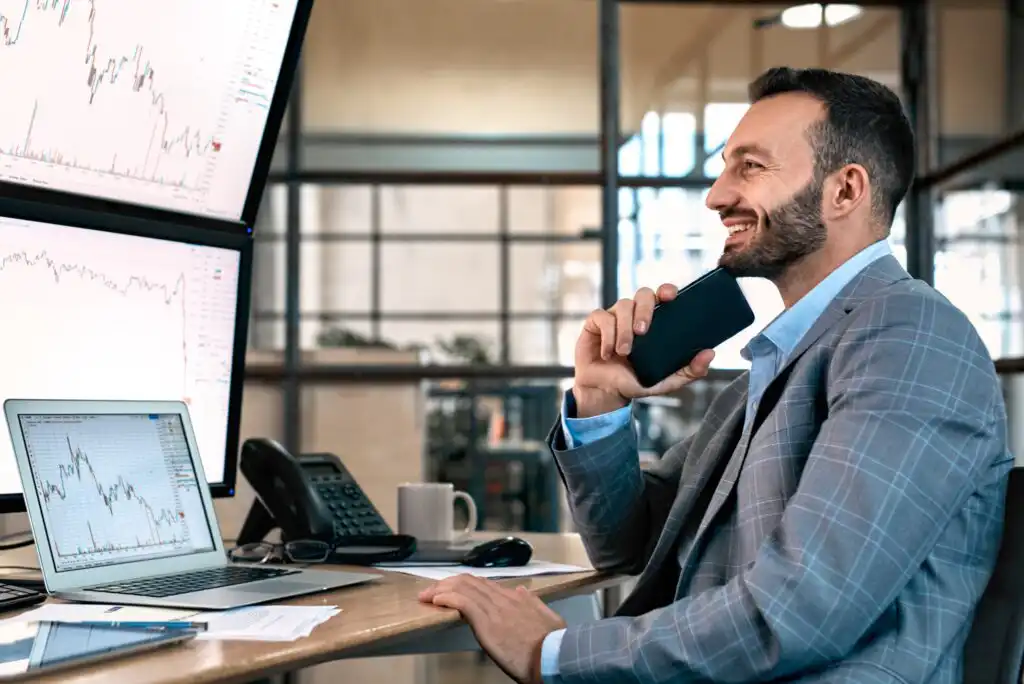 A man in a suit sits at a desk with multiple monitors and a laptop displaying stock charts, smiling and holding a smartphone, in a modern office setting.