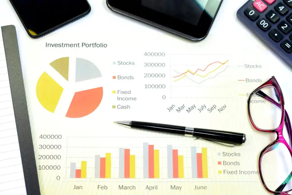 A financial document displays investment portfolio charts and graphs, including a pie chart, bar graphs, and a line graph. A pen, glasses, smartphone, and calculator are placed around the paper.
