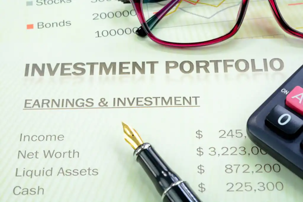 A close-up of a document titled “Investment Portfolio,” showing figures for income, net worth, liquid assets, and cash. A fountain pen, eyeglasses, and part of a calculator are also visible on the document.