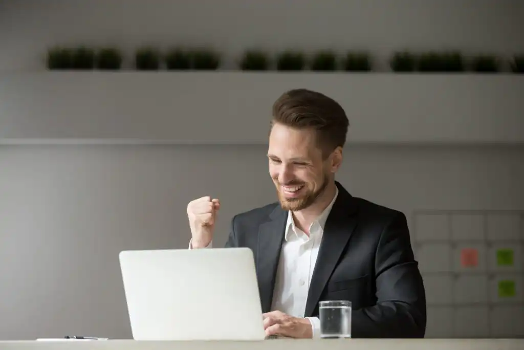 A man in a suit sits at a desk, smiling and raising his fist in excitement while looking at a laptop. There is a glass of water on the desk and green plants in the background.
