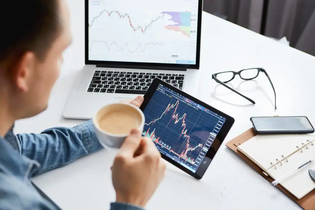 A person holds a coffee cup while looking at stock market charts on a tablet, with a laptop showing financial graphs, glasses, a smartphone, and a notebook on a white desk.