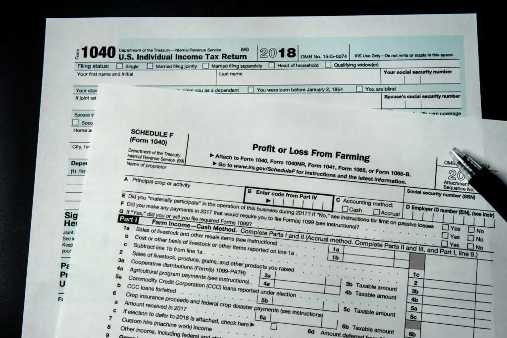 Two U.S. tax forms are shown: a 2018 Form 1040 Individual Income Tax Return and a Schedule F for Profit or Loss From Farming, with a black pen placed on top of the forms.
