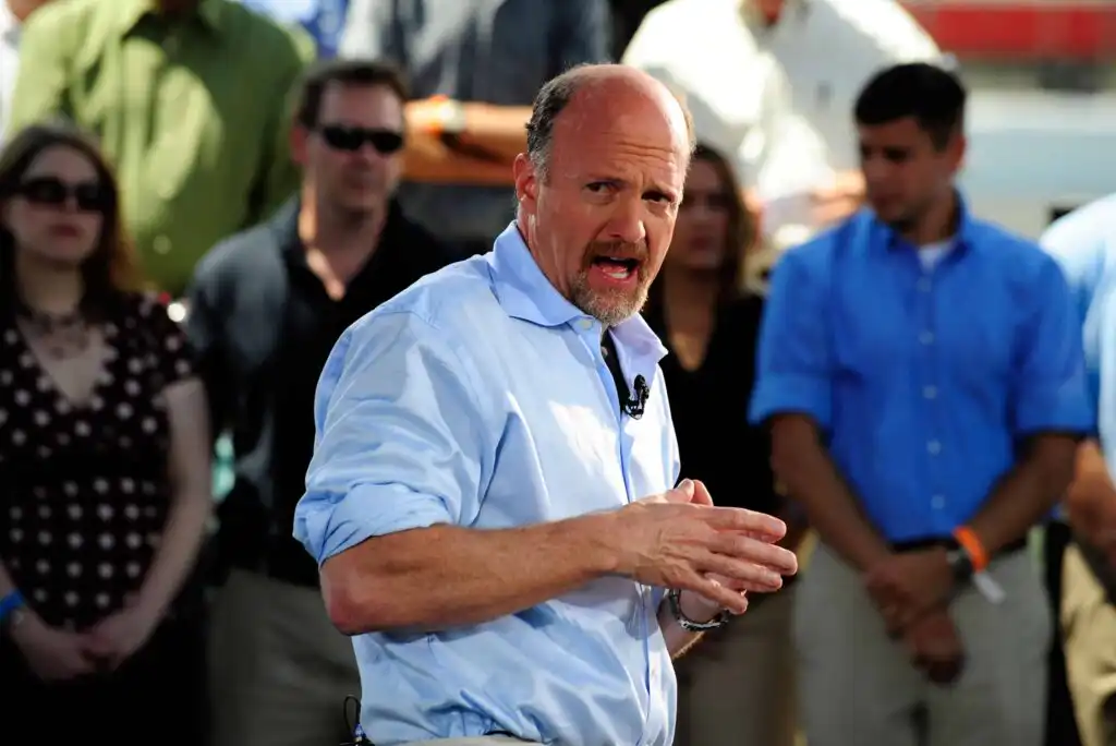 A man in a light blue shirt gestures while speaking to a group of people standing behind him outdoors. The crowd appears attentive, and the background is slightly out of focus.