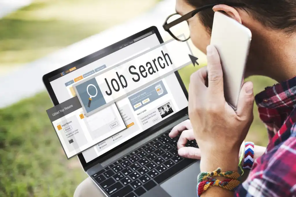 A person holding a smartphone looks at a laptop screen displaying a Job Search website, with search tools and options visible. The person wears colorful bracelets.