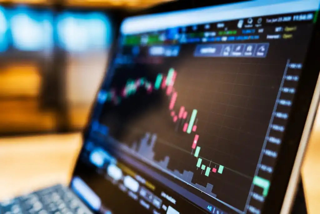 A close-up of a laptop screen displaying a colorful candlestick stock chart with various market indicators, representing financial data and trading activity. The background is blurred.