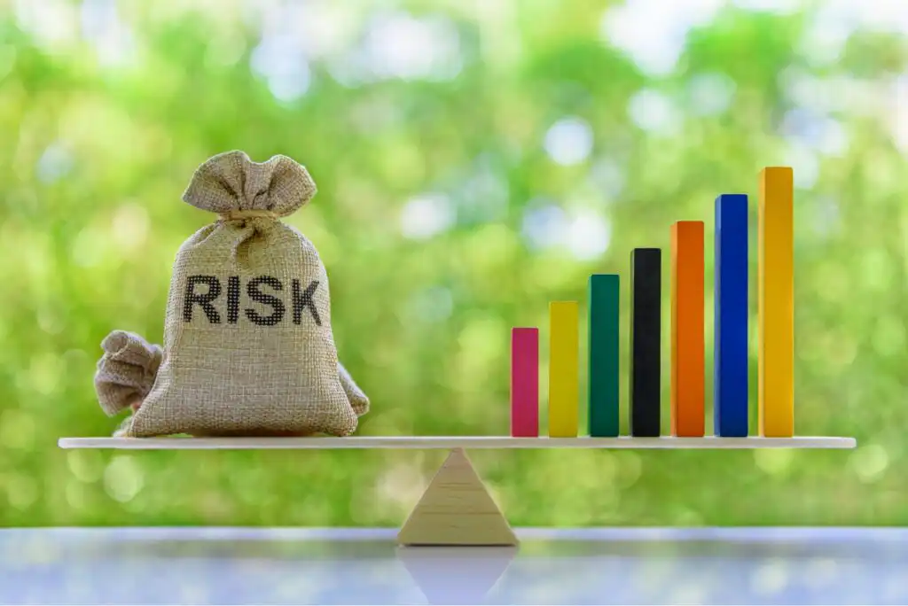 A burlap sack labeled RISK and colorful stacked bars are balanced on a wooden seesaw against a blurred green background.