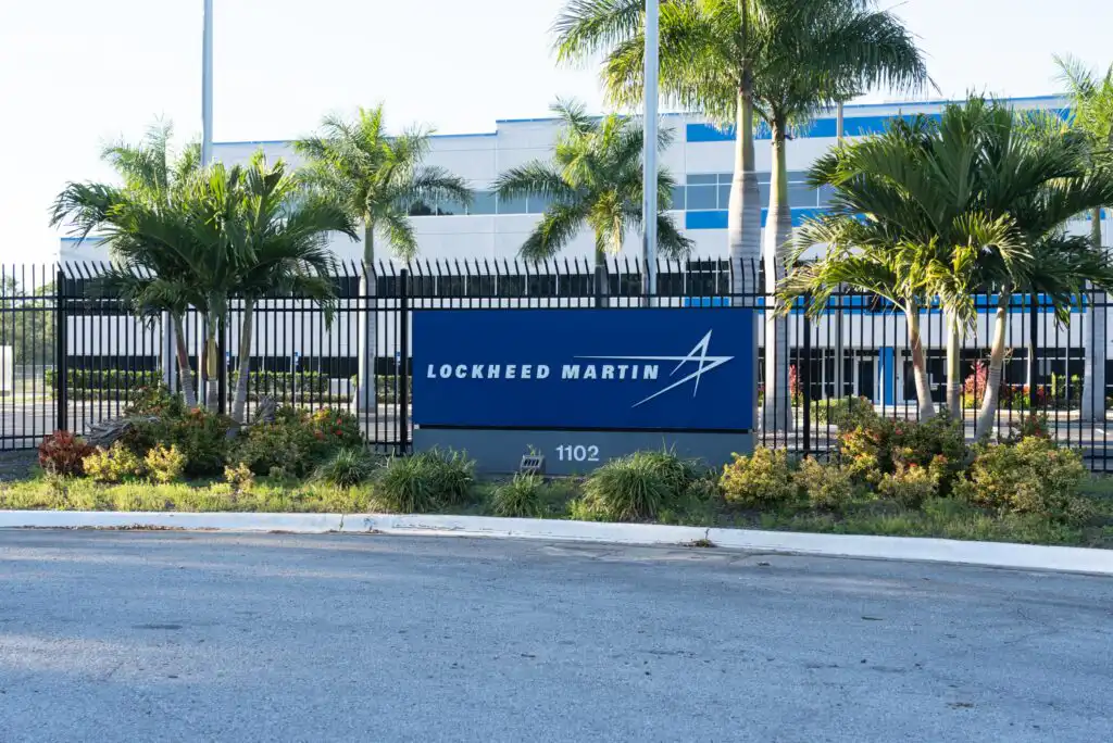 A Lockheed Martin sign with the company logo stands in front of a fenced office building, surrounded by palm trees and landscaping. The address 1102 is visible on the sign.