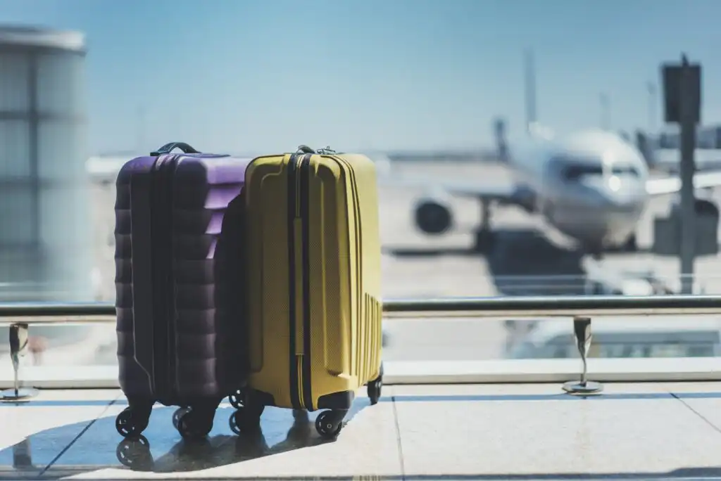 Two hard-shell suitcases, one purple and one yellow, stand side by side in an airport terminal with a blurry airplane visible through large windows in the background.