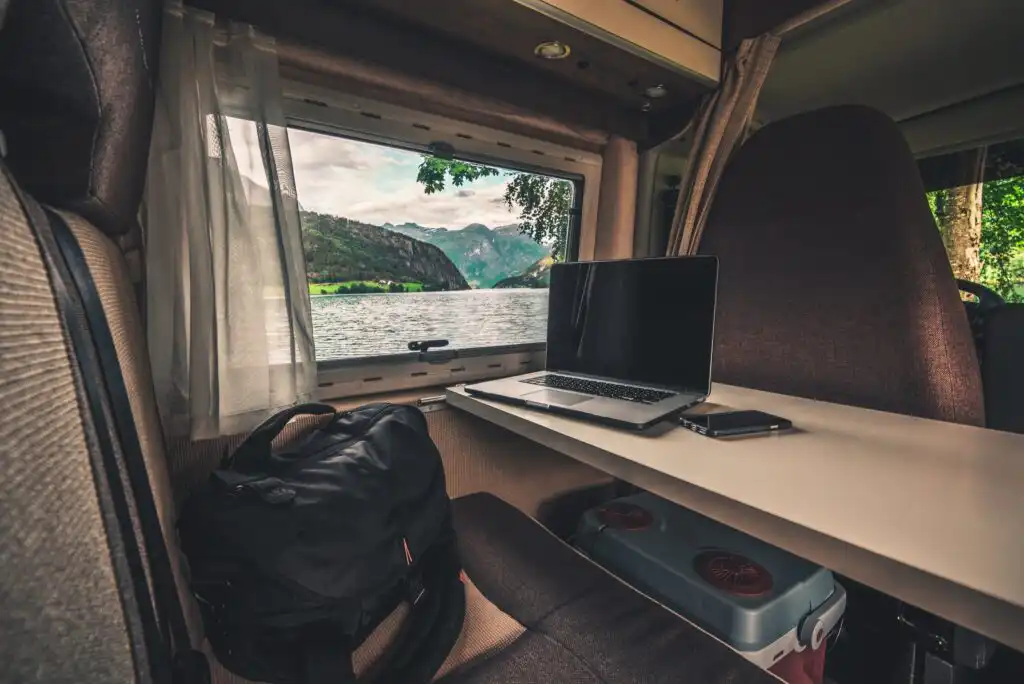 A workspace inside a camper van features a laptop, smartphone, and black backpack on a table, with a large window showing a scenic view of a lake, green hills, and mountains outside.