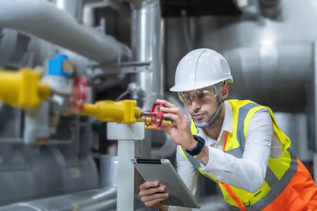 A male engineer wearing a white hard hat, safety glasses, and a yellow reflective vest inspects a valve in an industrial facility while holding a tablet. Industrial pipes and equipment are visible in the background.