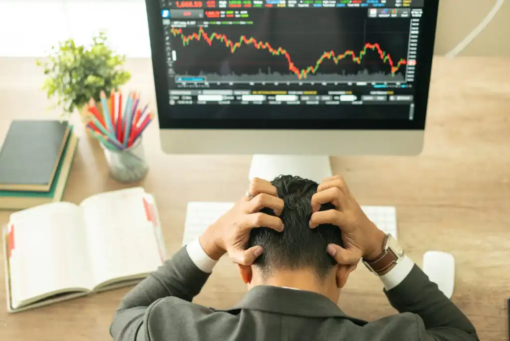 A person sits at a desk, clutching their head in frustration while looking at a computer screen displaying a stock market graph with a sharp decline. An open notebook, colored pencils, and a plant are on the desk.