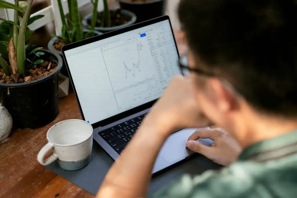 A person sits at a desk with a coffee cup, looking at a laptop screen displaying a financial chart or stock graph. Potted plants are in the background.