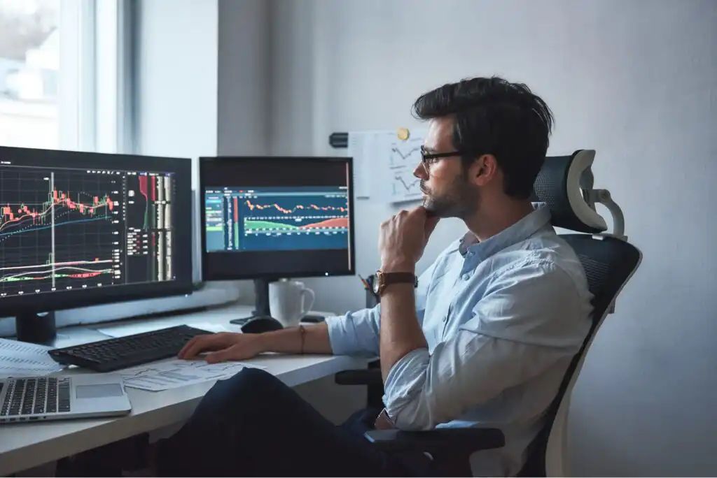 A man sits at a desk analyzing financial charts on multiple computer monitors, appearing focused and thoughtful, with papers and a coffee cup nearby in a modern office setting.