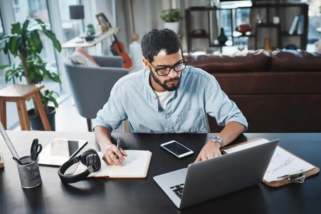 A man with glasses sits at a desk, working on a laptop while writing in a notebook. Headphones, a smartphone, and office supplies are on the desk. A living room with plants and shelves is visible in the background.