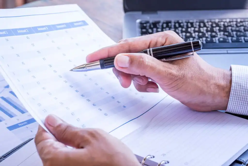 A person holds a pen and reviews a printed spreadsheet with numbers beside a laptop and an open notebook, suggesting planning or data analysis at a workspace.