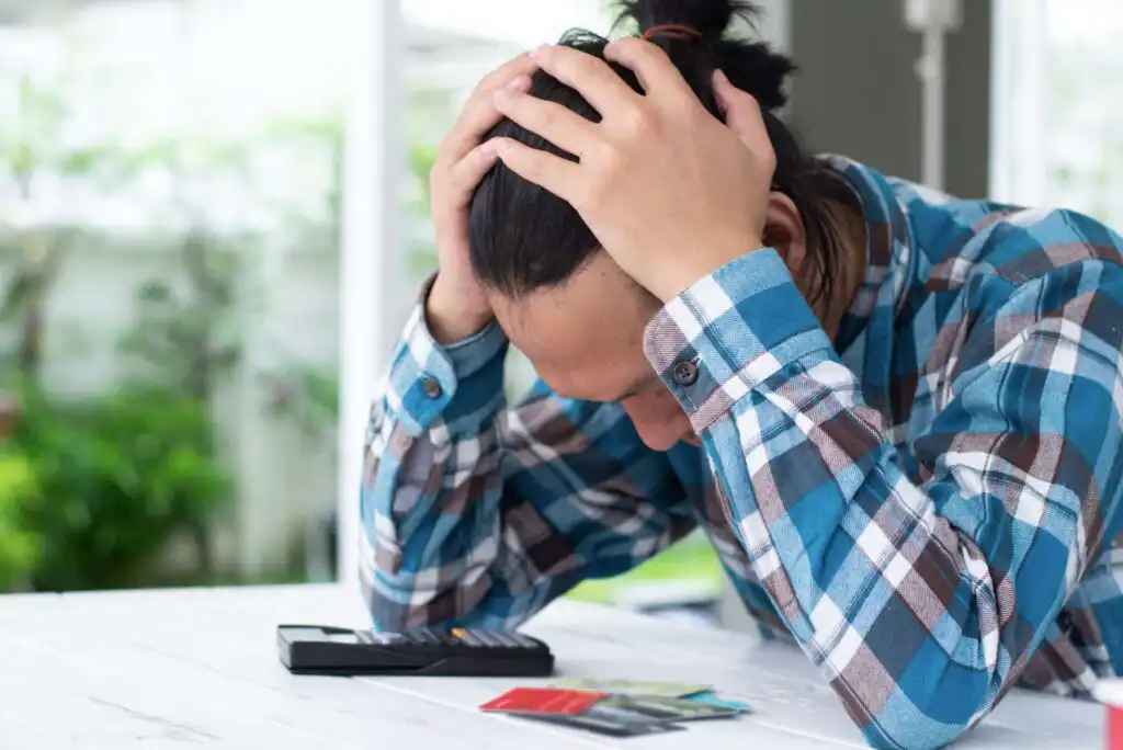 A person in a blue plaid shirt sits at a table with their head in their hands, looking stressed. A calculator and several credit cards are on the table in front of them.