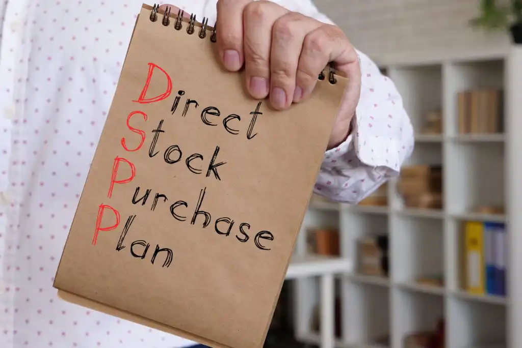A person holds a notebook with the words Direct Stock Purchase Plan written on it, with the initials DSPP highlighted in red. Shelves with books and boxes are blurred in the background.