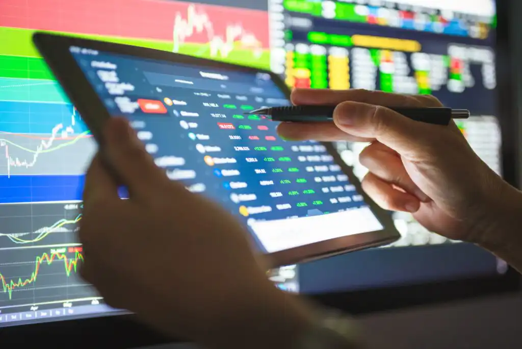 A person holds a tablet displaying stock market data and charts, using a pen to point at the screen. Colorful financial graphs and stock prices are visible on monitors in the background.