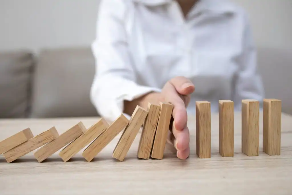 A person in a white shirt stops a line of falling wooden domino blocks with their hand, preventing the remaining blocks from toppling over on a light-colored table.