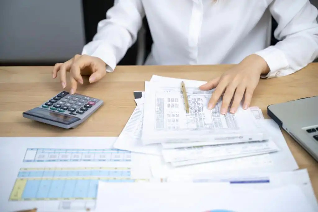 A person in a white shirt uses a calculator with one hand and reviews financial documents with the other, sitting at a desk covered with papers, spreadsheets, and a pen.