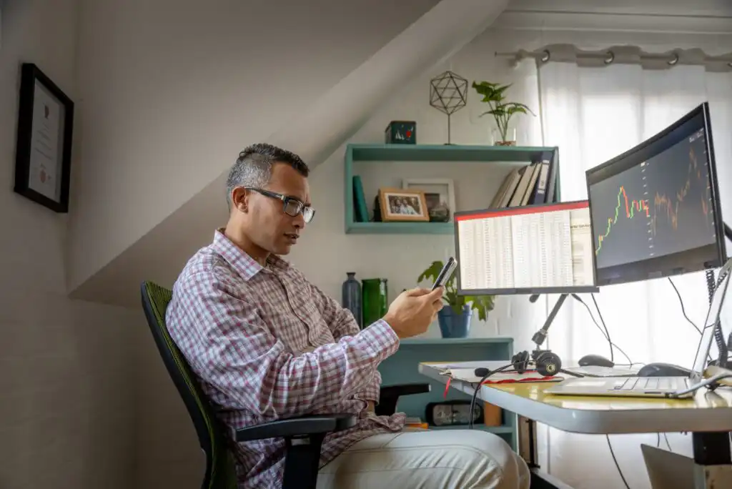 A man wearing glasses sits at a desk in a home office, looking at his phone. He is surrounded by computer monitors displaying charts and data, with shelves and plants in the background.