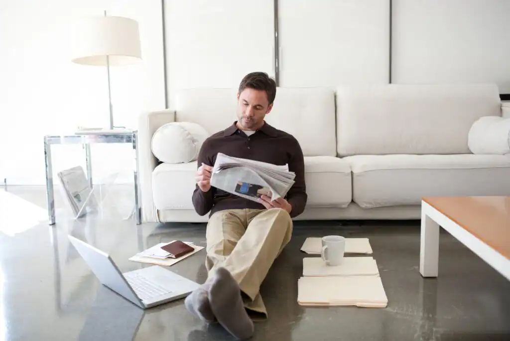 A man sits on the floor in front of a white sofa, reading a newspaper. A laptop, folders, a mug, and documents are spread around him. The room is bright and modern with a lamp and glass side table.