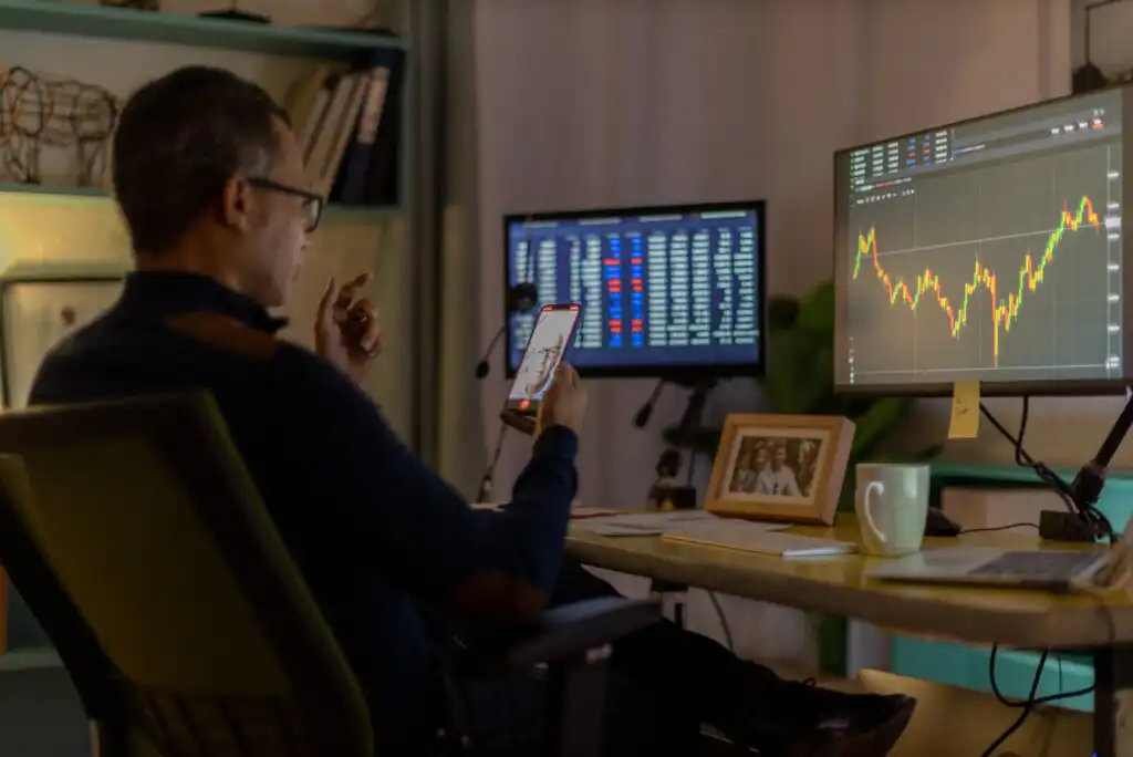 A person sits at a desk with two monitors displaying stock market data and charts, holding a smartphone. The desk has a coffee mug, framed photos, and office supplies. Shelves with books are in the background.