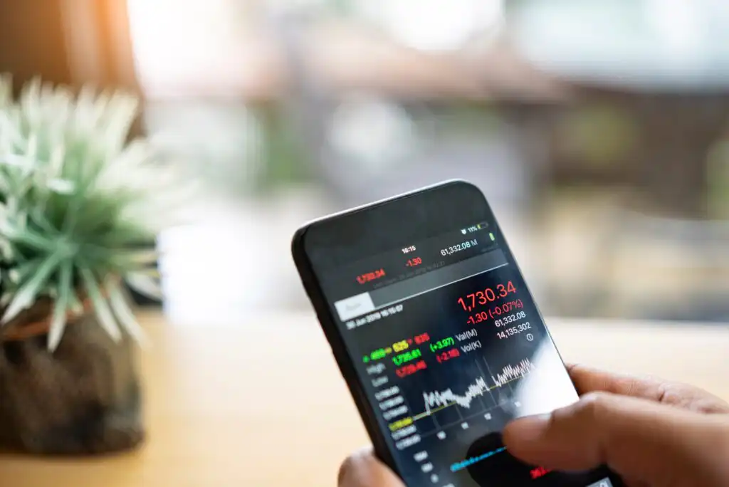 A hand holding a smartphone displaying a stock trading app with market data, red and green numbers, and a stock price chart. A blurred plant is visible in the background.