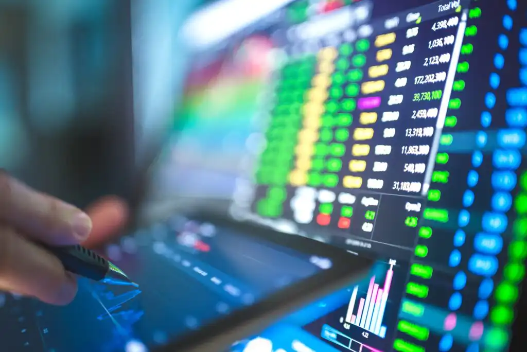 A hand holding a pen points at a digital tablet displaying stock charts, with colorful financial data and graphs visible on computer screens in the background.