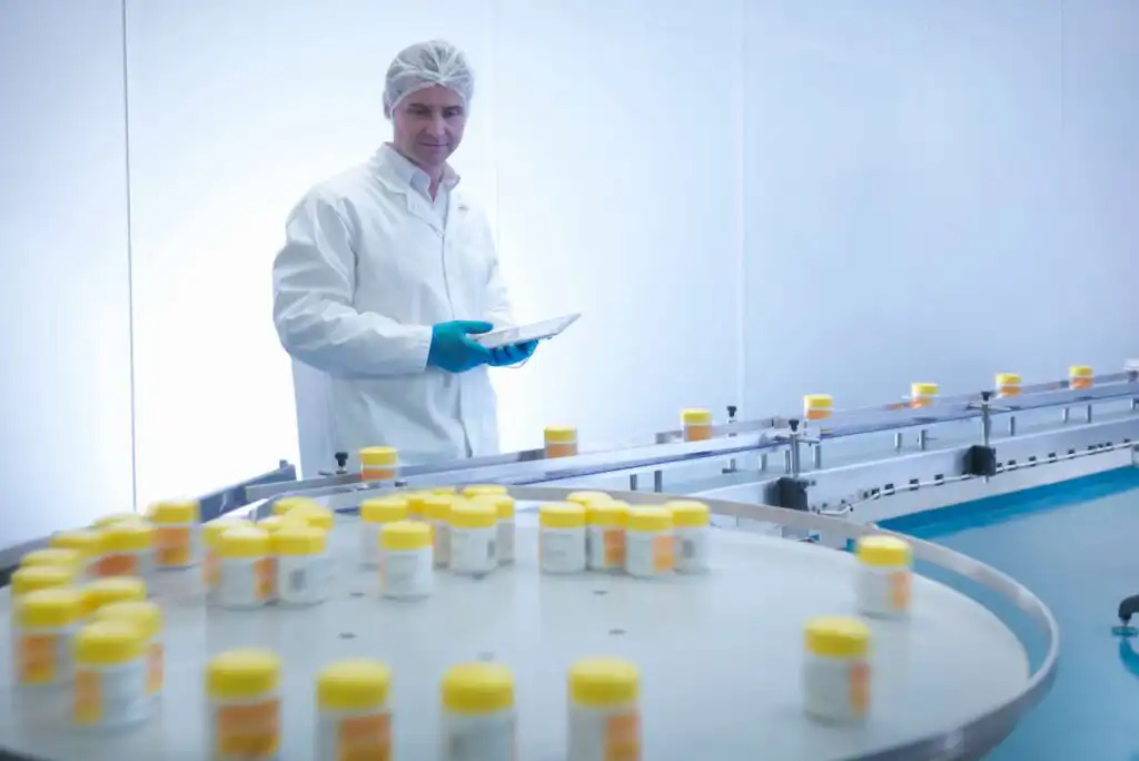 A person wearing a lab coat, hairnet, and gloves stands by a conveyor belt with multiple yellow-capped containers in a clean, modern laboratory or manufacturing facility.